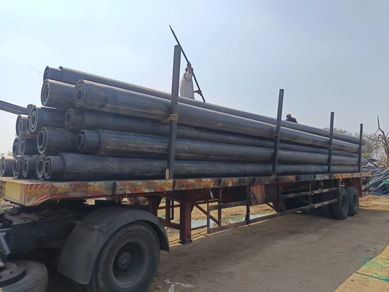 A flatbed trailer stacked with long black HDPE pipes, with crew securing the load on top under a clear sky.
