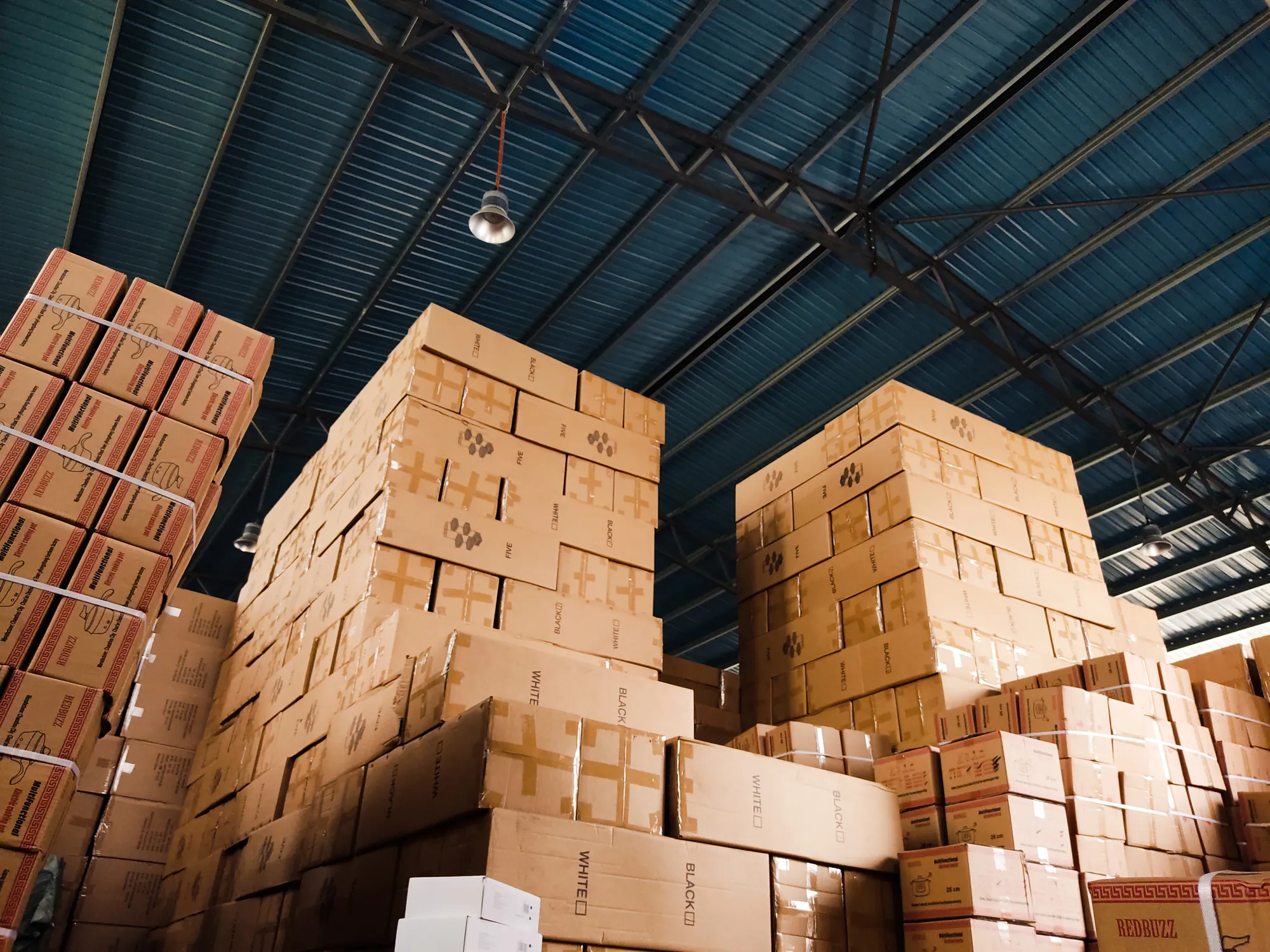 Cartons stacked high and staged under the steel roof trusses of a warehouse.